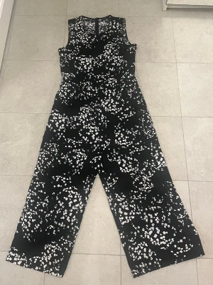 Svart blommig byxdress från Stockh LM - Snygg svart byxdress från Stockh LM i storlek 36 med vitt blommigt mönster. Modellen är ärmlös med rund hals och har en dragkedja i ryggen. Byxorna är vida och har en rak passform, perfekt för dig som gillar en trendig och bekväm look. 