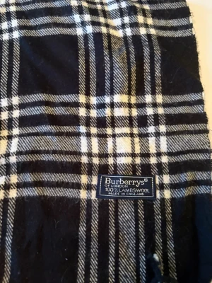 Rutig halsduk från Burberrys i ull - Snygg rutig halsduk från Burberrys, tillverkad i 100% lammull. Färgerna är svart, vit och grå, med klassiskt rutmönster och tvinnade fransar i ändarna. Perfekt för att ge din outfit en stilren touch under kyliga dagar.