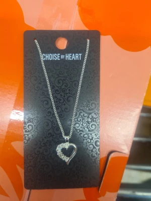 Halsband med hjärta från Choise by Heart - Snyggt silverfärgat halsband med ett hjärtformat hänge dekorerat med glittrande stenar. Perfekt accessoar för att lyfta din outfit och få uppmärksamheten.