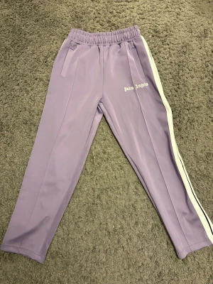 Lila Palm Angels trackpants - Snygga lila trackpants från Palm Angels med vit och svart rand längs sidorna. Byxorna har elastisk midja, sidofickor och Palm Angels-logga tryckt på framsidan. Byxorna är oanvända och riktigt fräscha. 