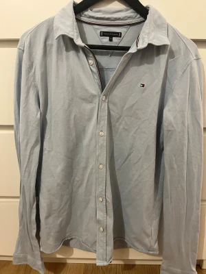 Ljusblå skjorta Tommy Hilfiger - Ljusblå skjorta från Tommy Hilfiger med klassisk krage och vita knappar framtill. Diskret Hilfiger-logga på bröstet och snygga detaljer med röd, vit och marinblå kant på insidan av kragen. Skjortan är långärmad och har en normal passform, perfekt för en clean och stilren look.