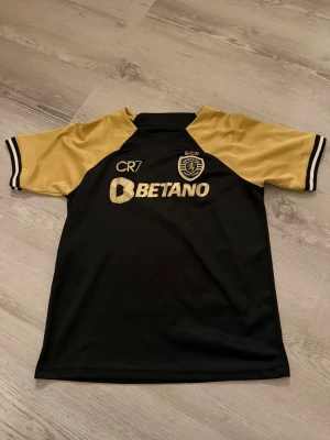 Sporting CP CR7 svart och guld fotbollströja - Snygg fotbollströja från Sporting CP med CR7-tryck och Betano sponsor på framsidan, Super Bock på ryggen. Svart kropp med guldiga ärmar och detaljer, samt vita och svarta ränder vid ärmslut. Tillverkad i lätt och ventilerande polyester.