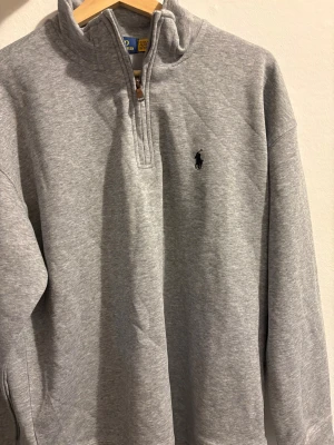 Grå half zip tröja från Polo Ralph Lauren - Snygg grå långärmad tröja från Polo Ralph Lauren med klassisk svart logga broderad på bröstet. Tröjan har en half zip-dragkedja vid halsen och är gjord i mjuk bomullsmix. Perfekt för chill dagar och enkel att matcha med jeans eller joggers.