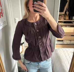 Mörklila kavaj med trekvartsärm - Snygg mörklila kavaj med rundad hals och stora knappar framtill. Jackan har en omlottliknande midja med rynkade detaljer och trekvartsärmar. Materialet känns som ull och ger en mjuk, lite lyxig vibe. Perfekt att styla med jeans för en cool look. Liten i storleken, sitter som en XS!