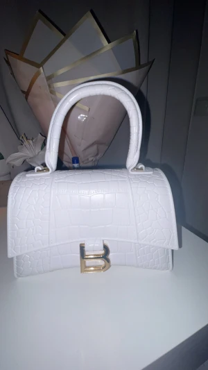 Vit handväska med guld telaj  - Snygg vit handväska  med krokopräglat mönster och guldfärgad B-detalj framtill. Väskan har ett stadigt handtag och medföljer en guldfärgad kedja för axelbärande. Perfekt storlek för det viktigaste och riktigt clean look.