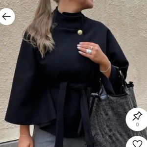 Svart kappa med bälte och knapp - Stilren svart kappa med hög krage, dekorativ guldig knapp vid halsen och knytskärp i midjan. Vida ärmar och elegant, minimalistisk design som ger en chic look. Perfekt för dig som vill ha en trendig och tidlös jacka. Från missay onsize som passar alla!