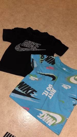 Nike t-shirt duo med tryck - Två snygga t-shirts från Nike. En svart med stort grafiskt Nike-tryck i vitt och en blå med färgglada Nike-loggor och texten 'Just Do It' i olika färger. Båda har rund hals och korta ärmar, perfekta för en sportig och avslappnad stil.