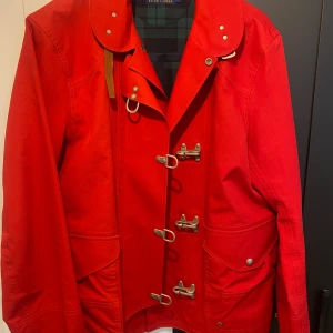 Röd jacka med spännen från raulph lauren nyskick, väldigt selsynt - Snygg röd jacka från Polo raulph lauren  med unika spännen framtill och stora fickor med knappar. Jackan har en klassisk krage och ett rutigt foder som ger en stilren look. Perfekt för höst och vår!