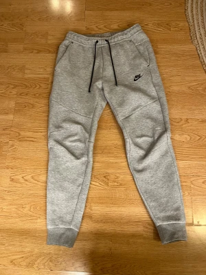 Nike tech byxor - Grå mjukisbyxor från Nike i storlek M med svarta snören i midjan och klassisk Nike-logga på vänster ben. Byxorna har ribbade muddar vid bensluten, två sidofickor och är tillverkade i mjukt bomullsmaterial. Perfekta för chill eller träning. Äkta!!