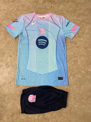 Barça Lamine Yamal #10 fotbollströja - Säljer en Barcelona fotbollströja med Lamine Yamal och nummer 10 på ryggen. Tröjan är ljusblå med rosa och mörkblå detaljer, kortärmad och tillverkad i Dri-Fit material. FC Barcelona-logga och Spotify-tryck på framsidan, UNHCR ACNUR på baksidan. Matchande mörkblå shorts med klubbmärke ingår.