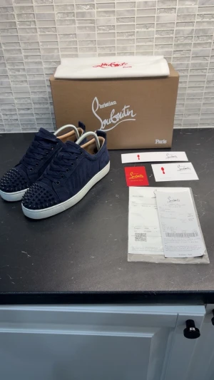 Christian Louboutin blå sneakers med nitar - Storlek 39 men sitter som 40-40,5.                  Säljer ett par mörkblå sneakers från Christian Louboutin med ikoniska röda sulor och coola nitar på tån. Skorna har snörning, rund tå och är tillverkade i mocka med vit platt sula. Perfekt för dig som vill sticka ut med lyxig streetstyle.