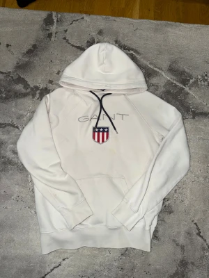 GANT Hoodie - Vit hoodie från GANT med broderat emblem i rött, blått och vitt på bröstet samt GANT-logga ovanför. Tröjan har huva med mörkblå snörning och en stor magficka. Tillverkad i mjuk bomull, perfekt för chill dagar. Klassisk och stilren design. Enstaka fläckar som går att få bort med hjälp av vanish.