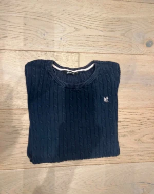 Mörkblå kabelstickad tröja Gina Tricot - Snygg mörkblå kabelstickad tröja från Gina Tricot med rund hals och diskret logga broderad på bröstet. Tröjan har långärmade smala ärmar och ribbade muddar vid hals, ärmslut och nederkant. Perfekt för dig som gillar klassisk stil.