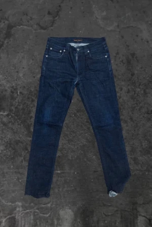 Mörkblå jeans från Nudie Jeans, 30/32 - Snygga mörkblå jeans från Nudie Jeans i klassisk straight fit. Jeansen har fem fickor, subtila sömmar på bakfickan och en svart läderpatch i midjan. Materialet är slitstarkt denim i bomull och passformen är normal. Perfekta för dig som gillar stilrena och tidlösa jeans.