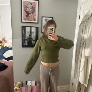 Olivgrön croppad stickad tröja - Säljer en olivgrön croppad stickad tröja med bred ribbad mudd i midjan och vid ärmslut. Tröjan har lång ärm och en klassisk rund halsringning. Perfekt för lager-på-lager och snygg till både jeans och kjol.