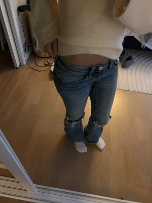Blå slitna jeans från Gina Tricot - Säljer ett par blå jeans från Gina Tricot med låg midja och raka ben. Jeansen har slitna detaljer och hål vid knäna för en cool, avslappnad vibe. Klassisk femficksmodell med kontrastsömmar och knappstängning framtill.