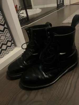Svarta kängor Dr. Martens - Säljer ett par Dr. Martens svarta kängor i blankt skinn med snörning och rund tå. De har en robust platt sula och dragflik baktill för enkel påtagning. 