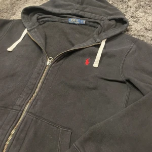 Svart hoodie från Polo Ralph Lauren - Svart hoodie från Polo Ralph Lauren med dragkedja och vit snörning i huvan. Klassisk röd logga broderad på bröstet. Mjuk insida och två fickor framtill. Storlek L. Tappat lite färg vid muddarna