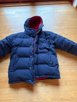 Mörkblå pufferjacka Tommy Jeans - Säljer en mörkblå pufferjacka från Tommy Jeans med röd insida och huva. Jackan har flera fickor med knappar, dragkedja och tryckknappar framtill samt justerbara axelband på insidan. Perfekt för kalla dagar. Hängslen inuti så du kan ”hänga av dig jackan” utan att lägga ifrån dig den. Använd fåtal gånger nyskick. 