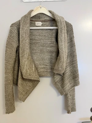 Beige stickad kofta från ONLY - Säljer en beige stickad kofta från ONLY med öppen front och avslappnad passform. Koftan har lång ärm och en snygg, omlottliknande drapering framtill. Perfekt att slänga över en t-shirt för en chill look.