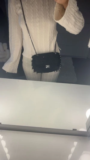 Svart quiltad axelväska från Rebecca Peterson - Snygg svart axelväska med glittrig quiltad yta och fransiga kanter. Väskan har en silverfärgad kedja som axelrem och ett stilrent metallspänne framtill. Man får plats med mycket i den trots att den ej är så stor 