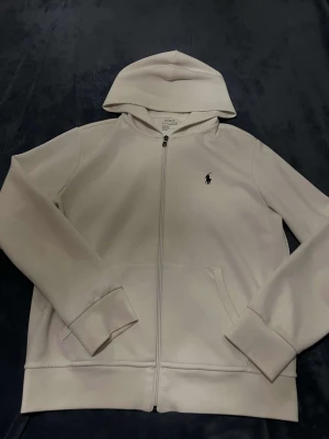 Vit hoodie från Polo Ralph Lauren - Snygg vit hoodie från Polo Ralph Lauren med dragkedja och huva. Klassisk logga broderad på bröstet i svart. Tröjan har två fickor framtill och är tillverkad i mjuk bomullsmix som är skön att ha på sig. Perfekt för chill dagar och enkel att matcha.