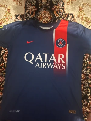 PSG Mbappé matchtröja Nike blå/röd 21/22 - Paris Saint-Germain fotbollströja med Mbappé 7 på ryggen. Djupblå med röd vertikal rand och vita detaljer, PSG-emblem på bröstet och Nike-logga. Qatar Airways sponsortryck framtill. Tillverkad i lätt och ventilerande polyester.