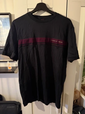 Svart Porsche BOSS t-shirt med vinröd detalj - Svart t-shirt från Porsche x Hugo Boss med vinröd och svart rand över bröstet. Klassisk rund hals och korta ärmar. Snyggt tryck med Porsche och BOSS på bröstet. Mjuk bomullskvalitet som känns skön mot huden.