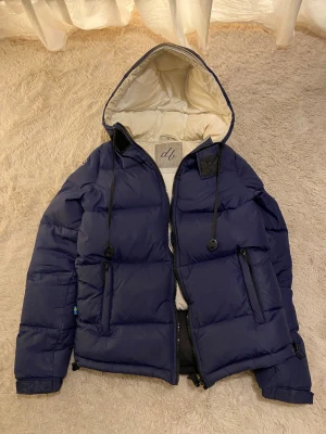 Blå vinterjacka med huva - Snygg blå vinterjacka från db med huva och vit insida. Jackan har broderad patch på ärmen, dragkedjor på fickorna och justerbar snörning vid huvan. Jackan är i S men passar nog bättre som XS. Den är ganska använd och har några fläckar på insidan vid det vita💞