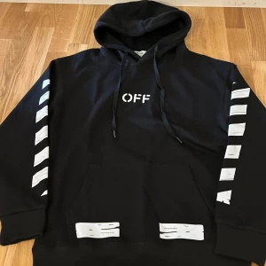 Svart Off-White hoodie med tryck - Svart hoodie från Off-White med vit grafisk print på både fram- och baksida samt ärmar. Klassisk huva med snörning och stor magficka. Snygga detaljer med texten 'OFF' framtill och 'SEEING THINGS' baktill. Perfekt för dig som gillar streetwear och statement-plagg.