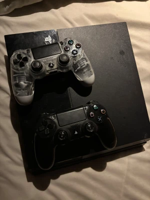 PlayStation 4 med två DualShock 4 kontroller - Säljer en PlayStation 4 med två original DualShock 4 kontroller (en svart och en transparent). Konsolen och kontrollerna är i bra skick med mindre yttre slitage. Perfekt för spelkvällar och multiplayer. Allt fungerar som det ska. Kan göra det billigare utan kontroller sladdar ingår