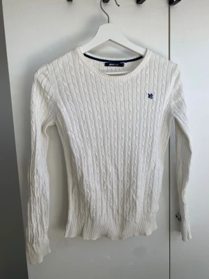 Vit kabelstickad tröja Gina Tricot - Snygg vit kabelstickad tröja från Gina Tricot med rund hals och ribbade muddar. Tröjan har ett broderat blått emblem på bröstet och är långärmad. Perfekt för dig som gillar klassisk och clean stil.