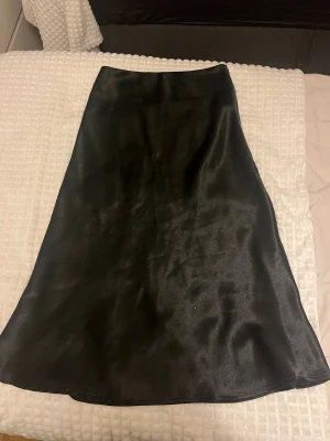 Svart långkjol i satin - En svart långkjol i glansigt satinmaterial med enkel och stilren design. Kjolen har en lätt utsvängd form som ger en snygg siluett och passar perfekt till olika toppar och skor. 