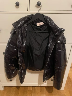 Svart pufferjacka från Nelly - Snygg svart pufferjacka från Nelly i glansigt syntetmaterial. Jackan har en stor huva, dragkedja framtill och quiltad design som ger en cool och modern look. Perfekt för kalla dagar och passar till streetwear-stilen.