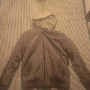 Brun bomberjacka med beige huva - Snygg brun bomberjacka från Kappahl men påminner om wrangler.Med beige huva. Jackan har dragkedja framtill, två fickor med lock och ribbade muddar vid ärmslut och nederkant. Materialet är glansigt och känns lätt, perfekt för dig som gillar streetwear och vill ha en chill look. Huvan kan tas av.