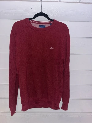 Vinröd stickad tröja från GANT - Snygg vinröd stickad tröja från GANT med diskret logga broderad på bröstet. Tröjan har rund halsringning och långa ärmar, perfekt för dig som gillar klassisk stil med en modern touch. Materialet är mjukt och strukturerat för extra komfort.