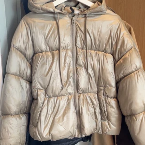 Beige pufferjacka med huva, stl M - Säljer en beige/guldig pufferjacka i storlek M med huva och dragkedja framtill. Jackan har en quiltad look och är perfekt för kyliga dagar. Den har snörning vid huvan och en lite oversized passform. Jag har ofta xs i storlek.