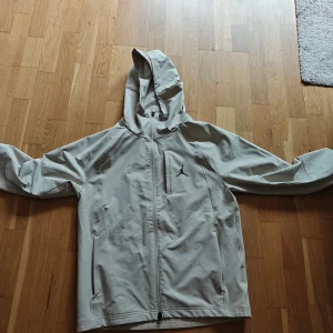 Beige vindjacka från Jordan med huva - Snygg beige vindjacka från Jordan med huva och dragkedja framtill. Jackan har en stilren look med diskret svart logga på bröstet och praktiska fickor med dragkedja. Perfekt för blåsiga dagar och enkel att matcha med streetwear.