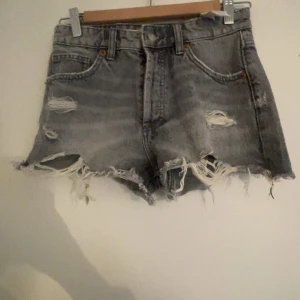 Grå jeansshorts med slitningar från Zara - Snygga grå jeansshorts från Zara med fransiga kanter. Shortsen har slitna detaljer framtill, klassiska fem fickor och knappar i gylfen. Perfekt för dig som gillar en edgy och avslappnad stil.