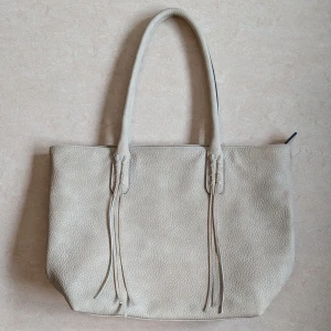 Beige axelväska med snördetaljer - Stilren beige axelväska från Åhléns med två långa handtag och snygga snördetaljer framtill. Väskan har dragkedja upptill, ett ytterfack med dragkedja och flera innerfack för smart förvaring. Perfekt för dig som vill ha en rymlig och klassisk väska.