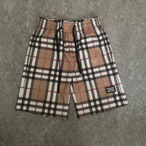 Burberry-rutiga shorts med snörning - Snygga shorts från Burberry med klassiskt rutig mönster i beige, svart, vitt och rött. De har elastisk midja med svart snörning och en patch med Burberry London England på benet. Perfekta för sommaren och ger en clean streetstyle-vibe.