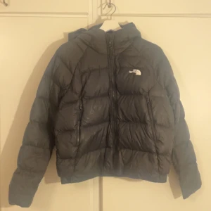 Svart dunjacka från The North Face - Säljer en svart dunjacka från The North Face i storlek L. Jackan har huva, två sidofickor med dragkedja och klassisk logga på bröstet. Puffad design med quiltade sektioner och hel dragkedja framtill. Perfekt för kalla dagar och riktigt snygg streetstil.
