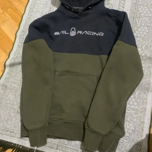 Sail Racing hoodie i marinblå och olivgrön - Snygg hoodie från Sail Racing i tvåfärgad design med marinblå upptill och olivgrön nedtill. Stor broderad logga på bröstet och mindre logga på huvan. Mjuk bomullsmix, känguruficka och justerbar huva med snörning. Perfekt för chill dagar.