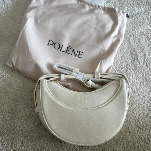 Vit axelväska från Polène i skinn - Säljer en superfin vit axelväska från Polène i strukturerat skinn. Väskan har en unik halvmåneform, justerbar axelrem och guldfärgade detaljer. Perfekt för dig som vill ha en stilren och trendig accessoar med exklusiv känsla.