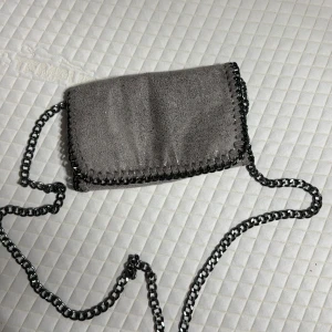 Grå axelväska med kedja - Snygg grå axelväska i glansig mocka-look med cool kedjerem och dekorativa sömmar längs kanterna. Väskan har magnetlås och ett innerfack med dragkedja. Perfekt för att lyfta din outfit med en edgy touch.