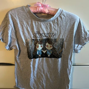 Grå The Shining t-shirt med tryck - Grå t-shirt med coolt The Shining-tryck framtill som visar de ikoniska tvillingarna i tecknad stil. Klassisk passform med rund hals och korta ärmar. Perfekt för dig som älskar skräckfilmer och vill sticka ut med en unik look.