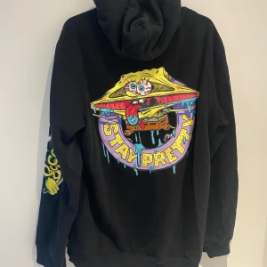 Svart hoodie med Spongebob tryck - Svart hoodie från H&M med färgglatt Spongebob-motiv på ryggen och mindre tryck fram. Har huva med snörning och stor känguruficka. Unik design med coola detaljer på ärmen och texten 'Stay Pretty' i lila och gult. Perfekt för dig som gillar streetwear och cartoon vibes.