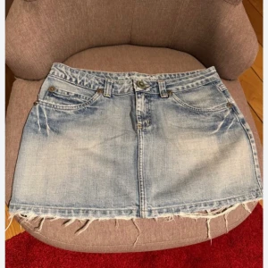 Ljusblå jeanskjol med fransig kant - Snygg ljusblå jeanskjol med slitna detaljer och fransig nederkant. Klassisk femficksmodell med bälteshällor och knappstängning framtill. Perfekt för en avslappnad och trendig look.