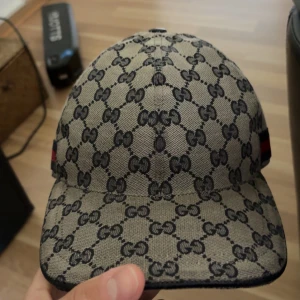 Gucci keps med GG-mönster och läderdetalj - Säljer en snygg keps från Gucci med klassiskt GG-mönster i beige och svart. Kepsen har justerbar rem i svart läder och en röd-blå rand på sidan. Tillverkad i canvas med insida i mjukt tyg. Perfekt för dig som vill sticka ut med en lyxig accessoar.