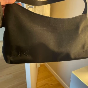 Svart axelväska från Donna Karan - Snygg svart axelväska från Donna Karan med stilren design och broderad DK-logga på framsidan. Väskan har en enkel, avlång form och är gjord i ett slätt syntetmaterial. 